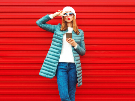 Fashion pretty woman blowing lips, using smartphone on a red background in the cityの写真素材