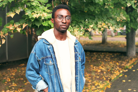 Fashion african man wearing jeans jacket, eyeglasses in autumn parkの写真素材