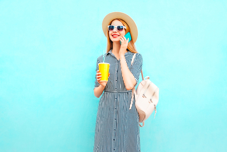 Fashion smiling woman is talking on smartphone holds cup of juice with backpack on colorful blue backgroundの写真素材
