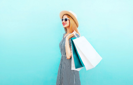 Fashion young woman walks with shopping bags in striped dress, summer round straw hat on blue backgroundの写真素材