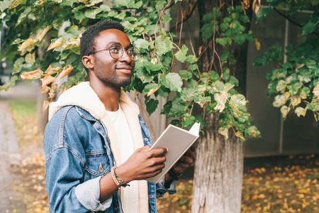 Happy smiling african man holds book, dreams in autumn city park near treeの写真素材