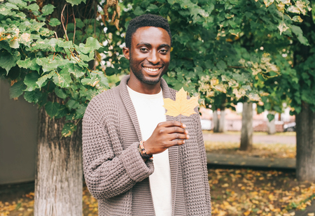 Happy smiling african man holds in hand yellow maple leaves in autumn day of city parkの写真素材