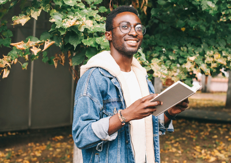 Happy young smiling african man holds book in autumn city parkの写真素材
