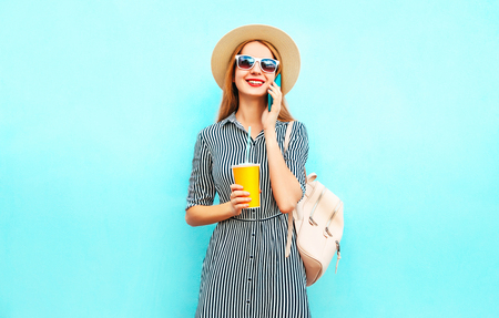 Portrait smiling woman is talking on smartphone holds cup of juice with backpack on colorful blue backgroundの写真素材