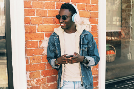 Fashion portrait cool african man with smartphone listening to music on city street, brick wall backgroundの写真素材