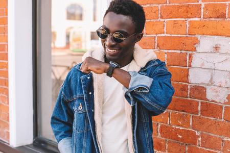 Fashion portrait smiling african man with smart watch using voice command recorder or takes calling on city street, brick wall backgroundの写真素材
