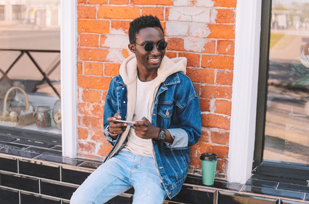 Fashion portrait smiling african man with smartphone sits on city street, brick wall backgroundの写真素材