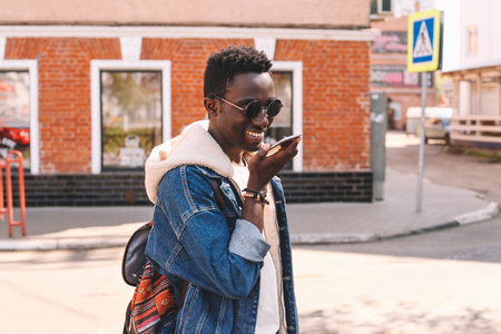 Fashion portrait smiling african man with smartphone using voice command recorder or calling walks on city streetの写真素材