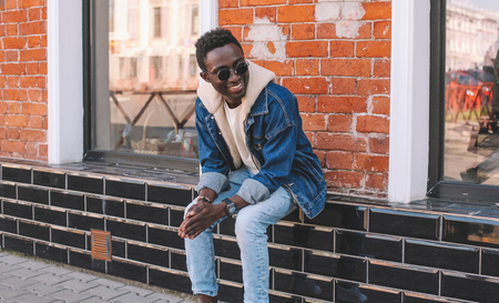 Fashion smiling african man wearing jeans jacket sits on city street, brick wall backgroundの写真素材