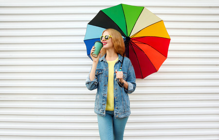 Happy smiling woman drinks coffee, holds colorful umbrella on white wall backgroundの写真素材