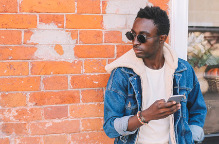 Fashion african man holds smartphone on city street, brick wall backgroundの写真素材