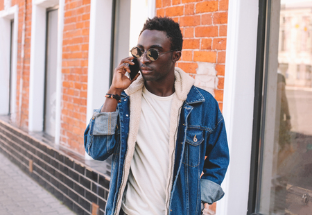 Fashion african man talking on smartphone walks on city street, brick wall backgroundの写真素材
