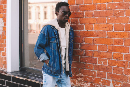 Fashion african man wearing jeans jacket poses on city street, brick wall backgroundの写真素材
