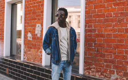 Fashion african man wearing jeans jacket poses on city street, brick wall backgroundの写真素材