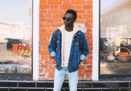 Fashion african man wearing jeans jacket, backpack poses on city street, brick wall backgroundの写真素材
