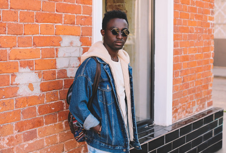 Fashion african man wearing jeans jacket, backpack poses on city street, brick wall backgroundの写真素材
