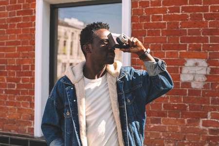 Fashion african man with vintage film camera taking picture on city street, brick wall backgroundの写真素材