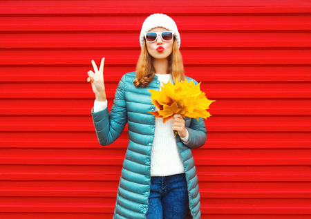 Fashion autumn woman holds yellow maple leaves, blows red lips, sends an air kiss on background in cityの写真素材