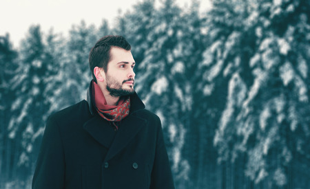 Fashion bearded brunette man walking in winter day, looks away on snowy forest trees backgroundの写真素材