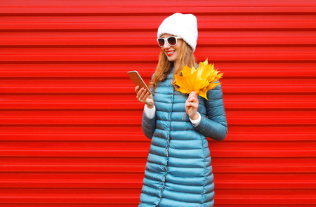Fashion autumn smiling woman using smartphone holds yellow maple leaves in hands on a red background in the cityの写真素材