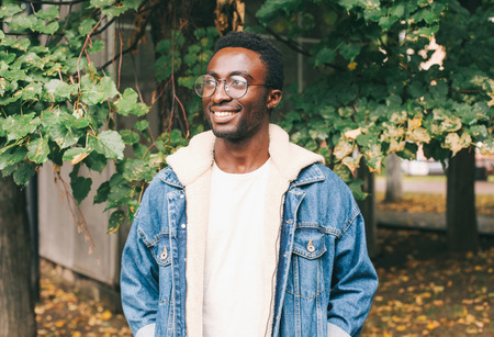 Fashion portrait smiling african man wearing jeans jacket, eyeglasses in autumn city parkの写真素材