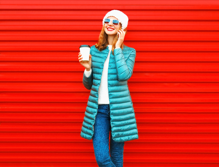 Fashion smiling woman talks on a smartphone holds coffee cup on a red background in the cityの写真素材