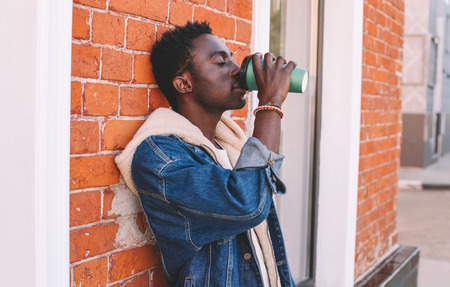 Break time! Fashion african man drinks coffee on city street, brick wall backgroundの写真素材