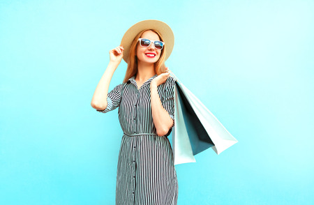 Fashion smiling woman with shopping bags in striped dress, round straw hat on blue backgroundの写真素材