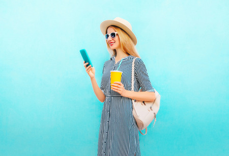 Fashion smiling woman is using smartphone holds cup of juice with backpack on blue backgroundの写真素材