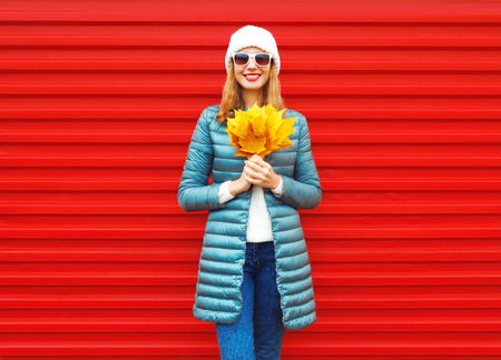 Fashion autumn smiling woman holds yellow maple leaves in her hands on a red background in the cityの写真素材