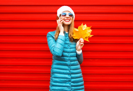 Fashion autumn smiling woman talks on a smartphone holds yellow maple leaves in hands on a red background in the cityの写真素材