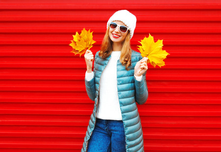 Fashion autumn smiling woman holds yellow maple leaves in hands on a red background in the cityの写真素材