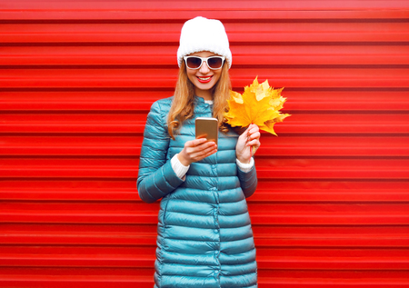 Fashion autumn smiling woman using smartphone holds yellow maple leaves on red background in the cityの写真素材