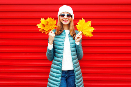 Fashion autumn smiling woman holds a yellow maple leaves in hands on red background in the cityの写真素材