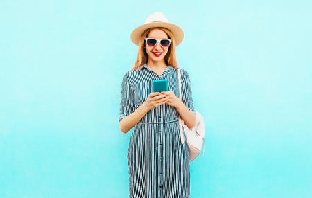 Fashion smiling woman is using smartphone wearing striped dress, backpack, straw hat on blue backgroundの写真素材