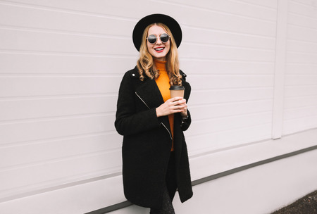 Break time! Stylish smiling young woman with coffee cup walking in city on gray wall backgroundの写真素材