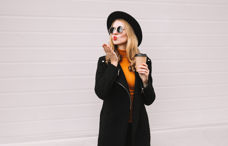 Beautiful woman blowing red lips sending sweet air kiss with coffee cup, wearing black clothes, jacket coat, round hat, gray wall backgroundの写真素材