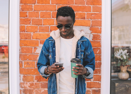 Fashion african man with smartphone, holds coffee cup on city street, brick wall backgroundの写真素材