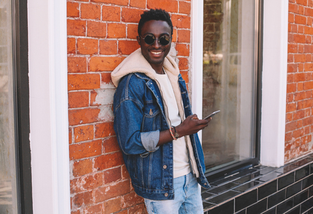 Fashion cool smiling african man with smartphone on city street, brick wall backgroundの写真素材