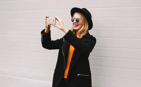 Fashion, technology and people concept - stylish smiling woman taking selfie picture by smartphone, elegant female model in black jacket coat, round hat in city on gray wall backgroundの写真素材