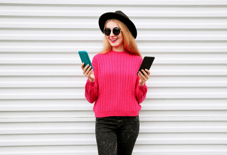 Cheerful cool girl holding two smartphone and looking at them in city on white wall backgroundの写真素材