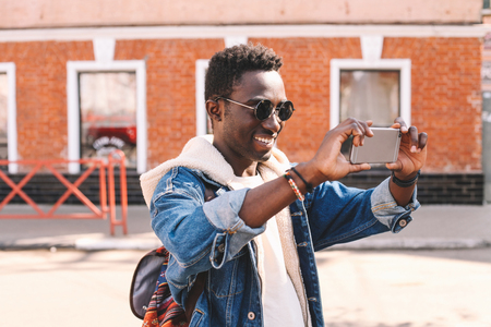 Cheerful smiling african man taking selfie picture by smartphone walking on city streetの写真素材