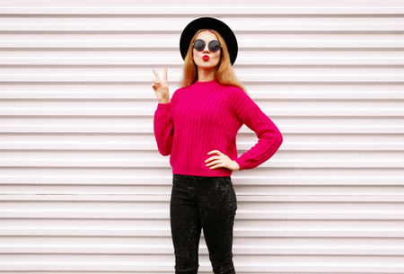 cool cheerful girl blowing red lips sending sweet air kiss in colorful pink knitted sweater, black round hat on white wall backgroundの写真素材