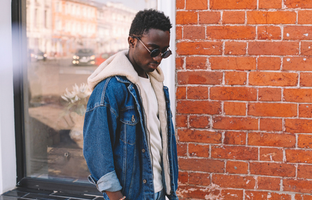 Fashion african man wearing jeans jacket walking on city street, brick textured wall backgroundの写真素材