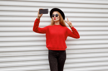 Fashionable young woman blowing red lips sends sweet air kiss taking selfie picture by phone in knitted sweater on white wall backgroundの写真素材