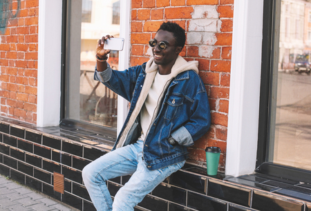 Happy smiling african man taking selfie picture by smartphone sitting with coffee cup on city streetの写真素材