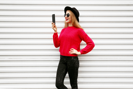 Happy smiling woman holding smartphone in colorful pink knitted sweater on city white wall backgroundの写真素材
