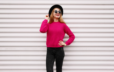 Stylish happy smiling woman in colorful pink knitted sweater, black round hat on white wall backgroundの写真素材