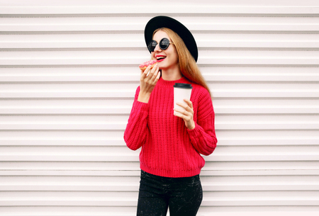 Portrait cheerful young woman eating sweet donut and holding coffee cup on city street white wall backgroundの写真素材