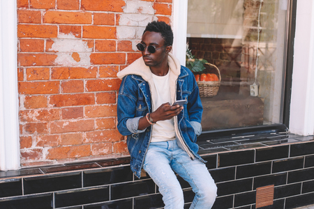 Fashion portrait cool urban african man with smartphone on city street, looking away, brick wall backgroundの写真素材